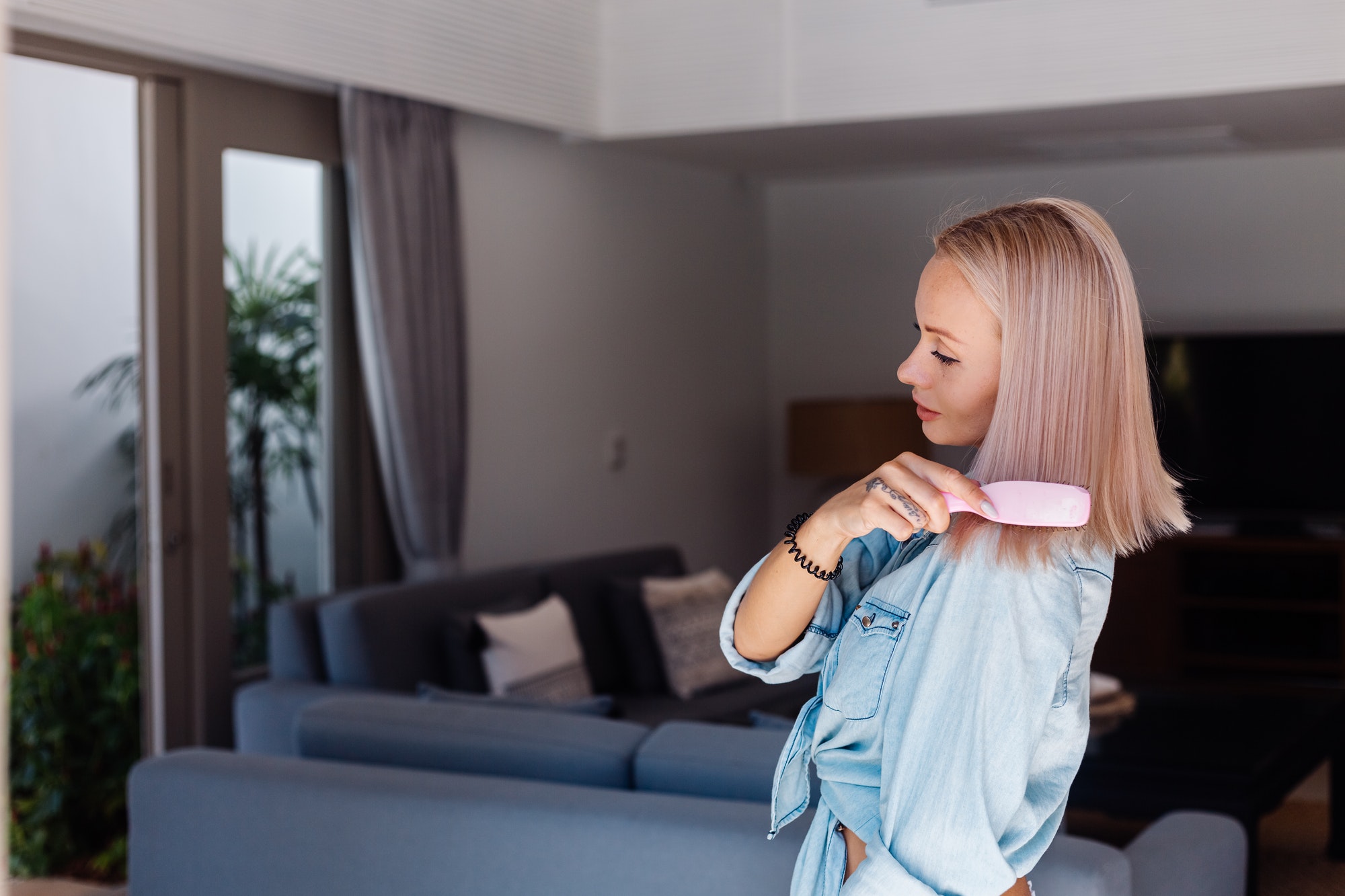 short hair woman in denim jacket brushing hair with pink hairbrush in living room